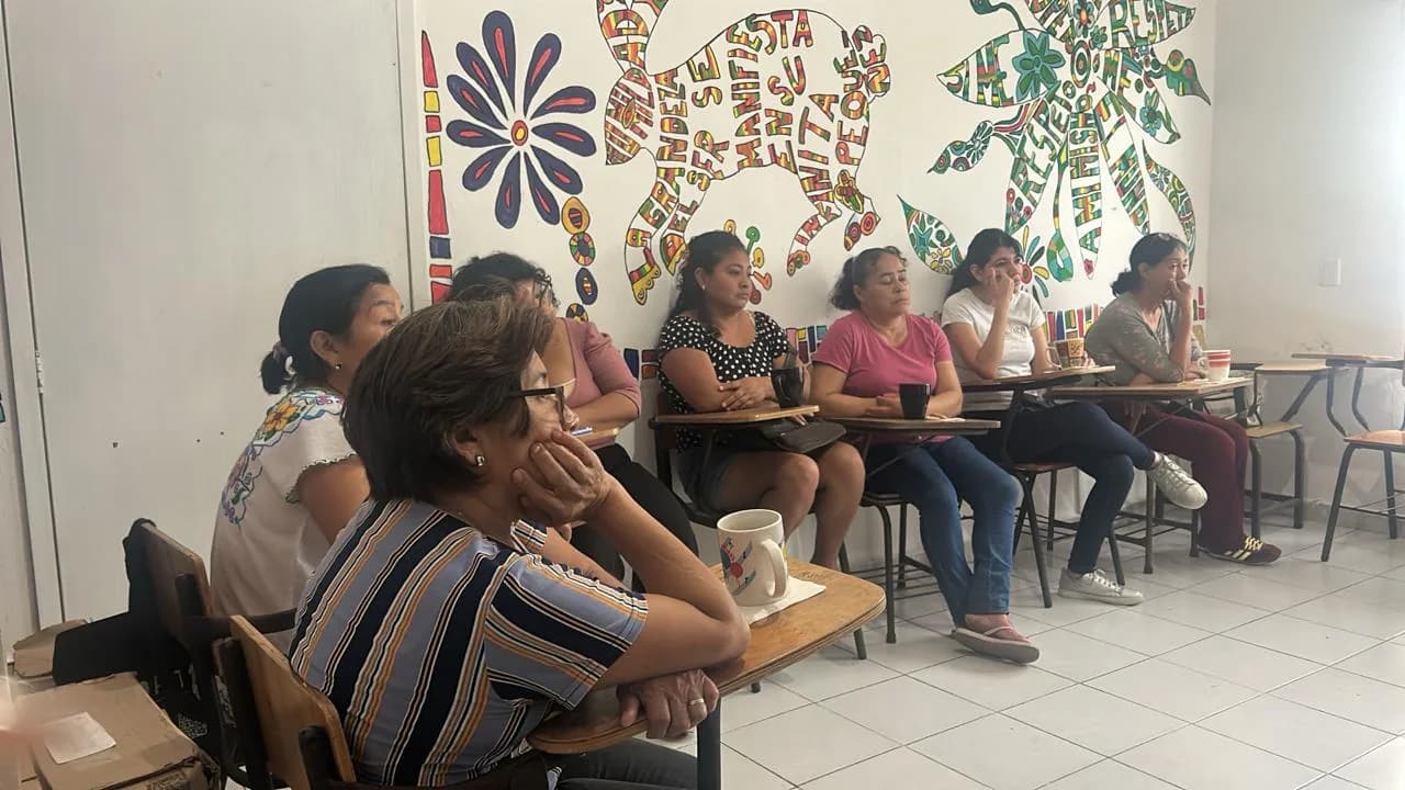 Mujeres atentas a una charla, sentadas en pupitres frente a un mural de colores.