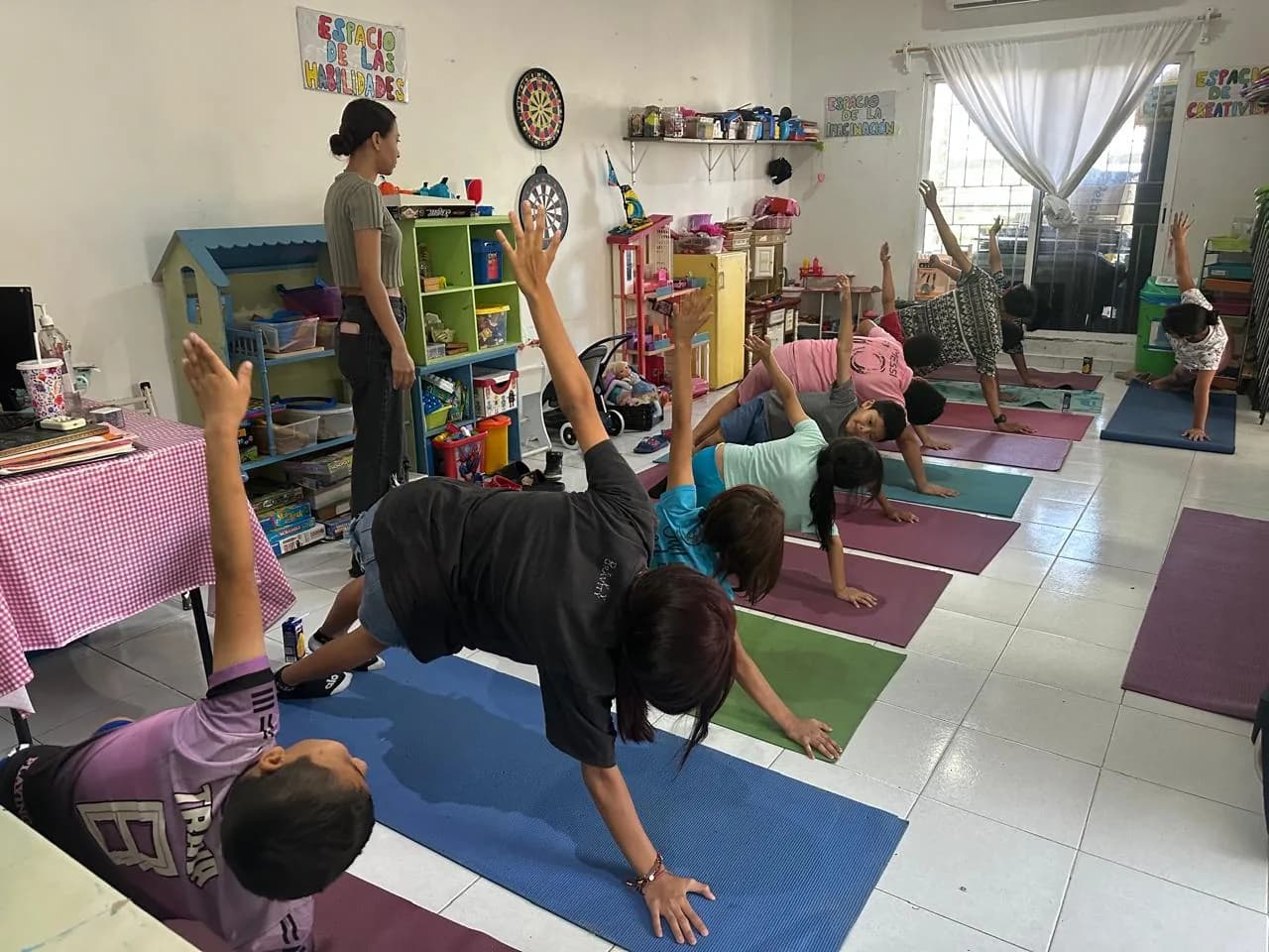 Niñas y niños hacen posturas de yoga sobre tapetes mientras una tallerista los acompaña.