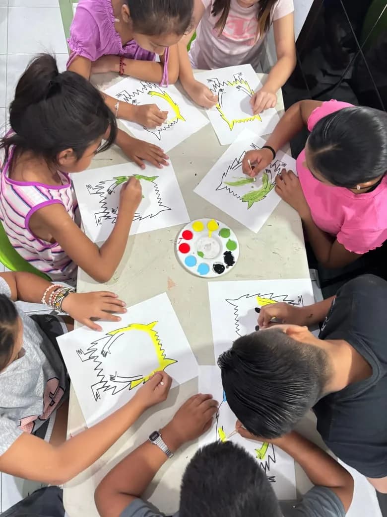 Vista desde arriba de una mesa donde varias niñas y niños pintan el mismo dibujo con acuarela.