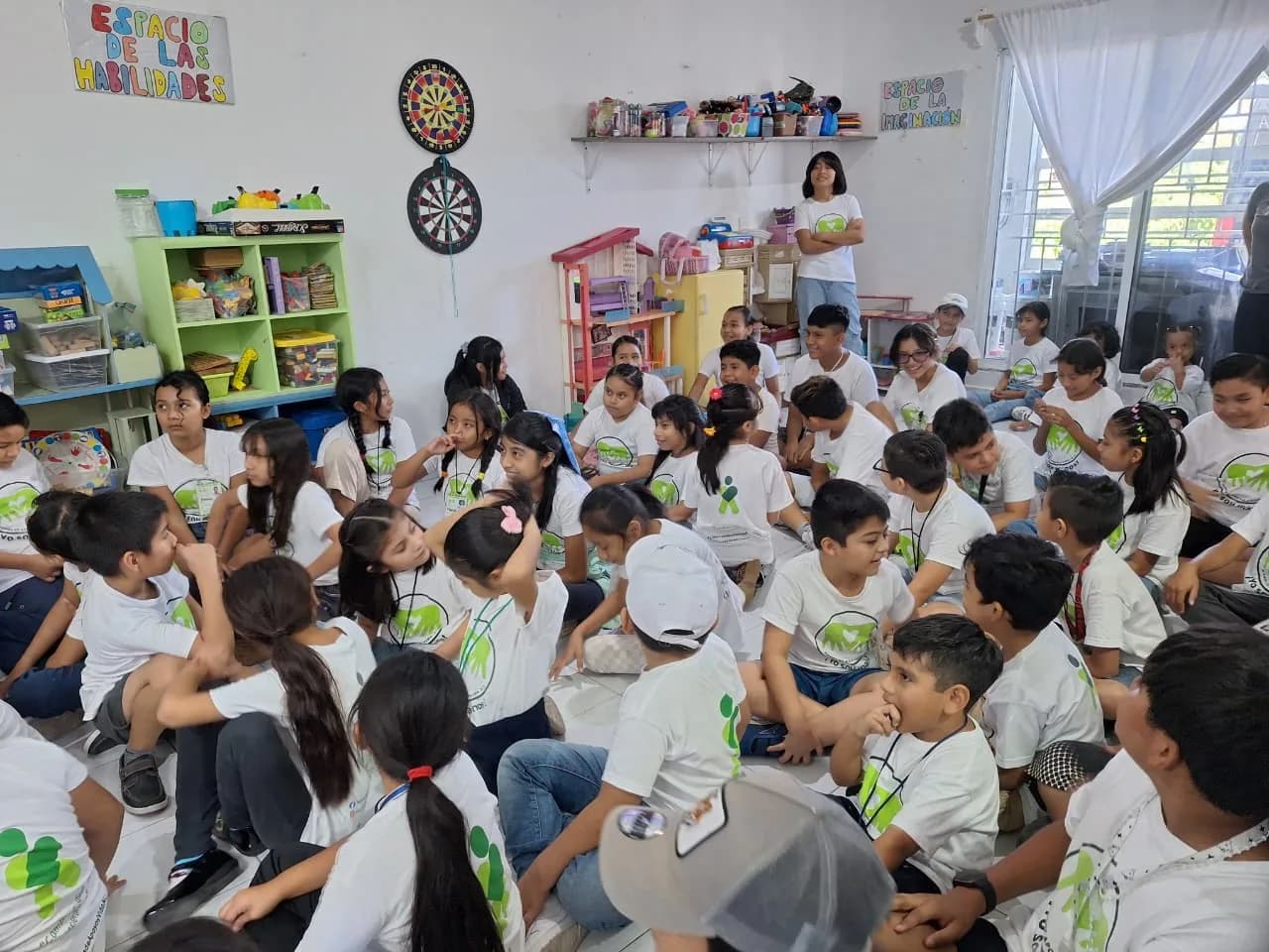 Un grupo numeroso de niñas y niños con playeras blancas escucha a una tallerista en la ludoteca.