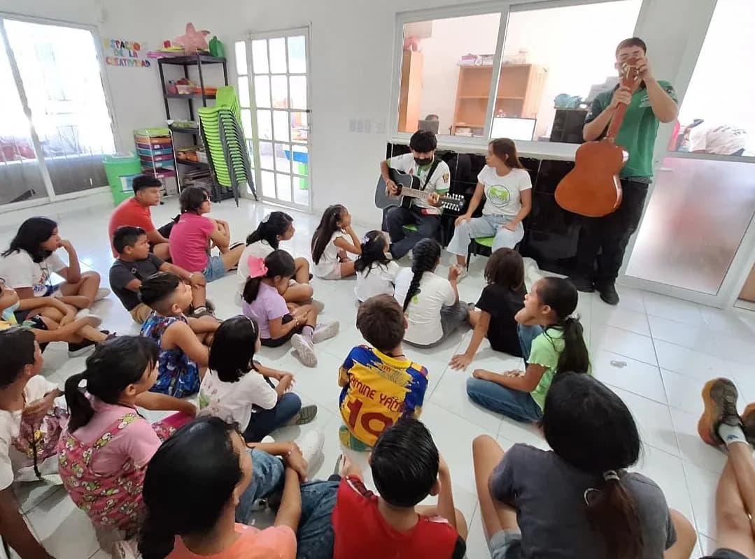 Niñas y niños sentados en el piso escuchan a dos jóvenes que tocan la guitarra.