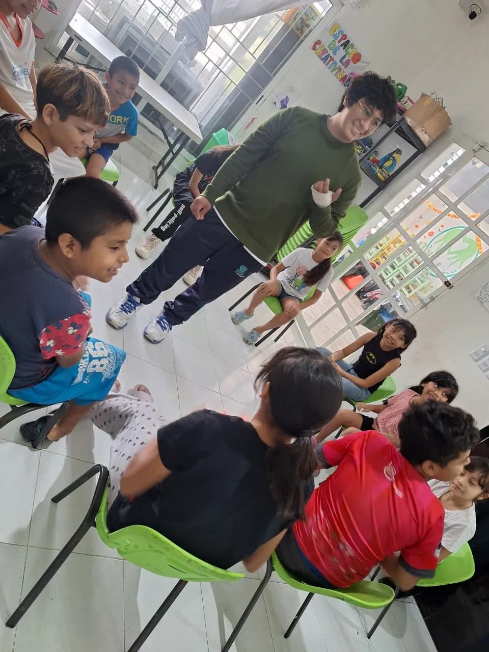 Un joven en servicio social dirige un juego con niñas y niños sentados en círculo.