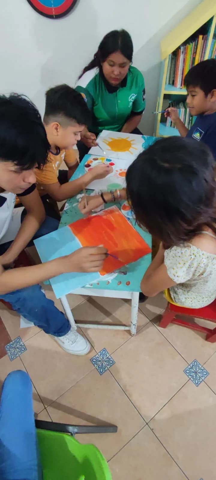 Una joven en prácticas pinta con témpera junto a tres niños sobre una mesa pequeña.