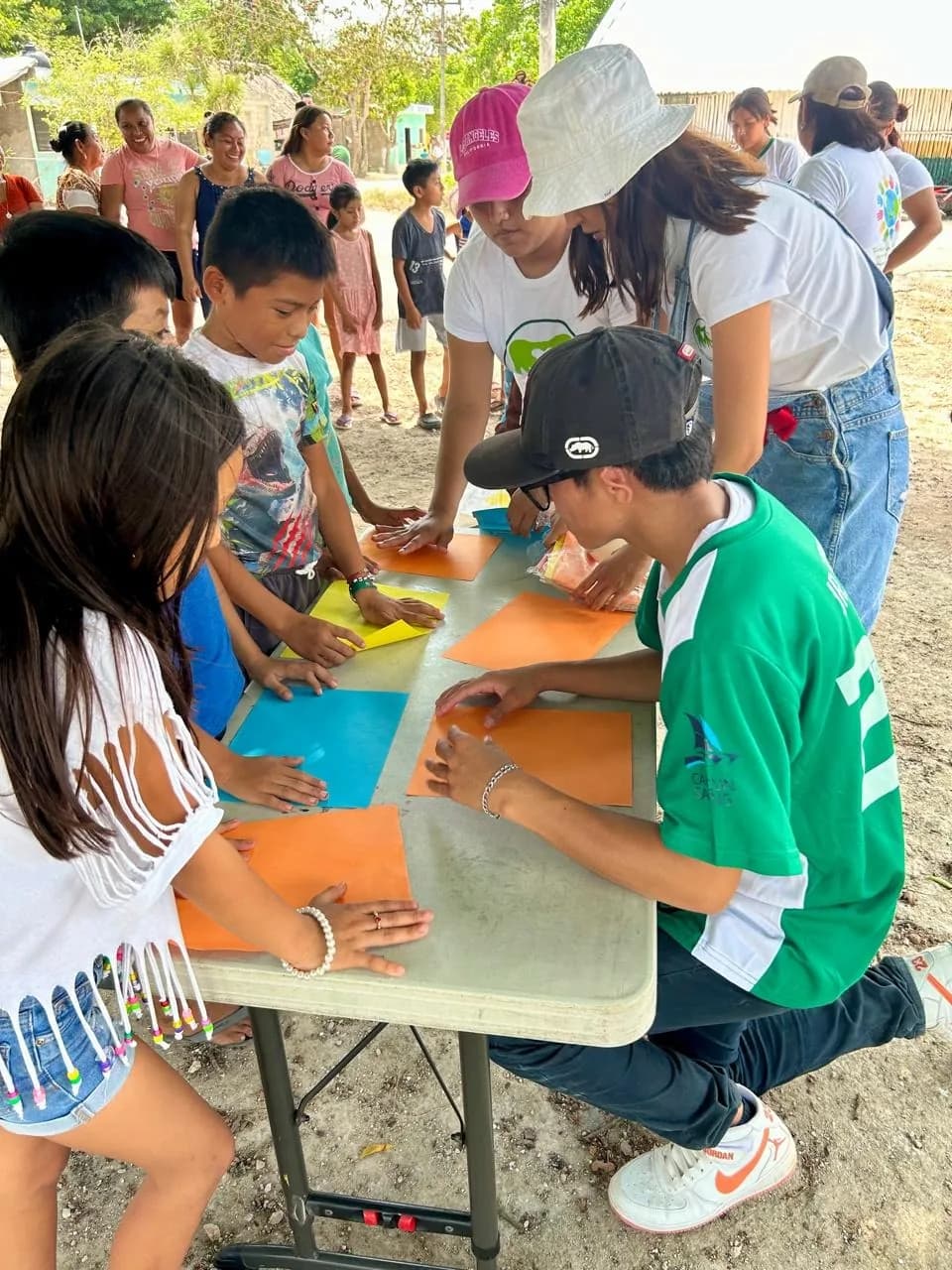 Voluntarias y voluntarios reparten material de manualidades a niñas y niños al aire libre.