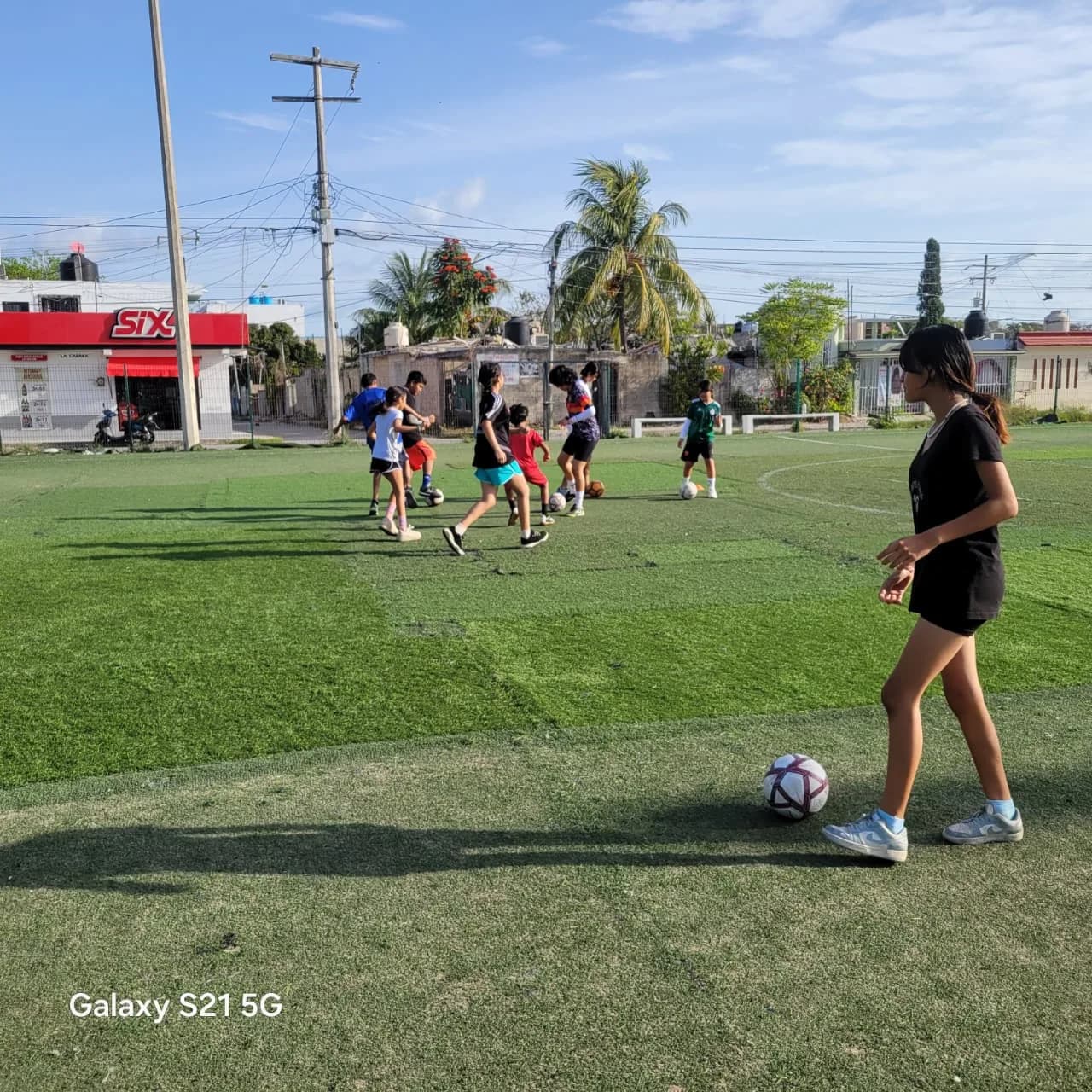 Niñas y niños entrenan con balón en una cancha de pasto sintético al atardecer.